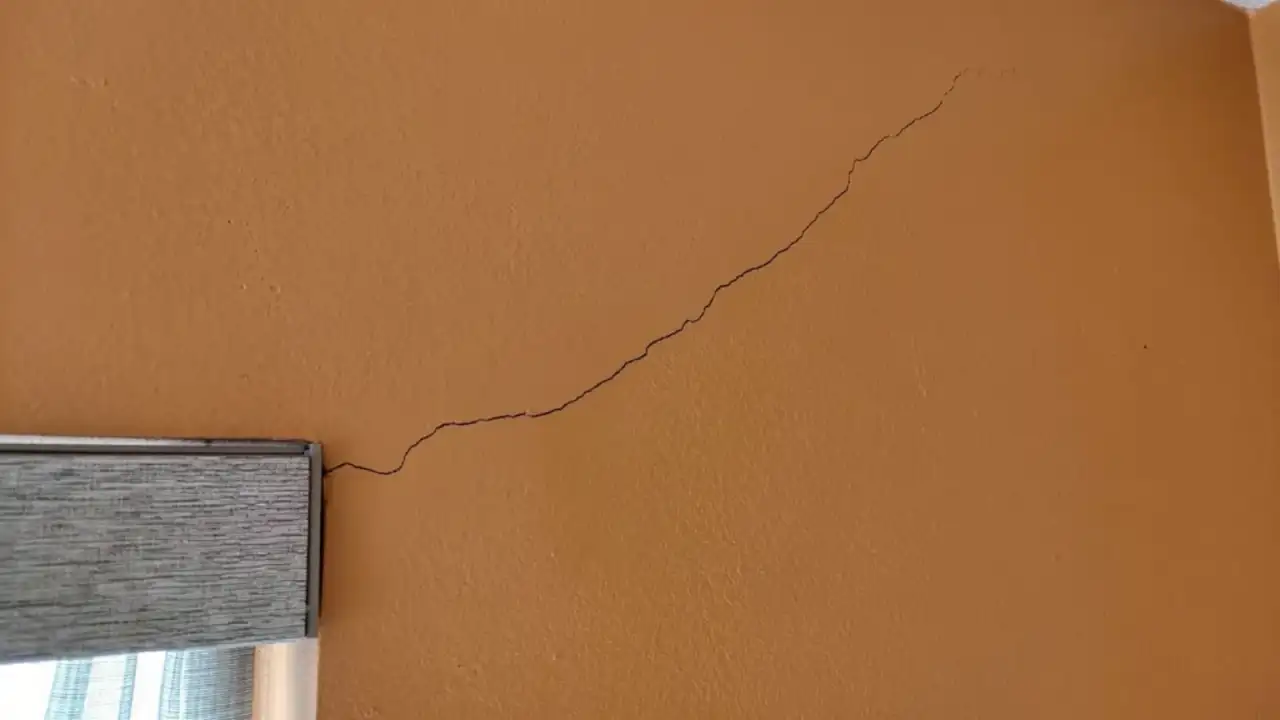 A hairline stress crack running diagonally across a painted interior wall from the head of a window casing up toward the wall-and-ceiling corner
