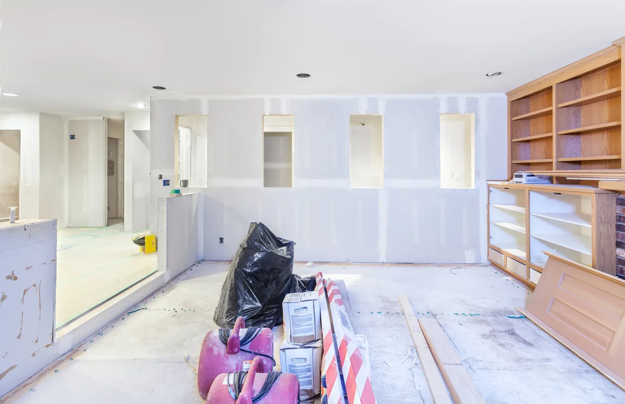Interior room with taped and mudded drywall walls, built-in wood shelving along one wall, recessed lights, and stacked materials on a bare floor before finishing