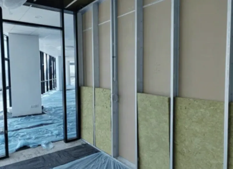 Commercial office fit-out in progress: steel-stud partition framing with drywall partly hung and mineral-wool insulation, floor protection down