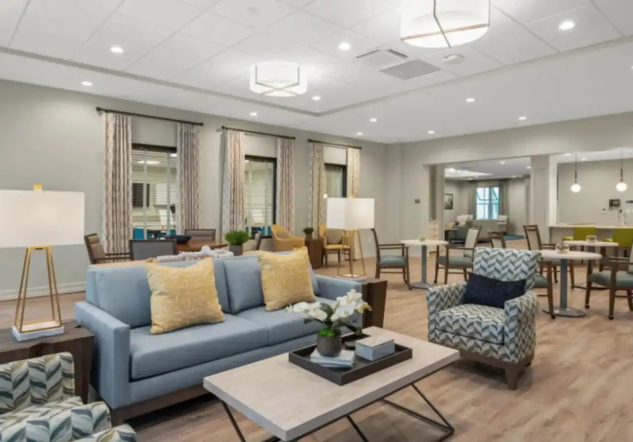 Senior-living community lounge with finished painted drywall walls, a suspended ceiling with recessed and pendant lighting, curtained windows, and upholstered sofas and armchairs around a coffee table